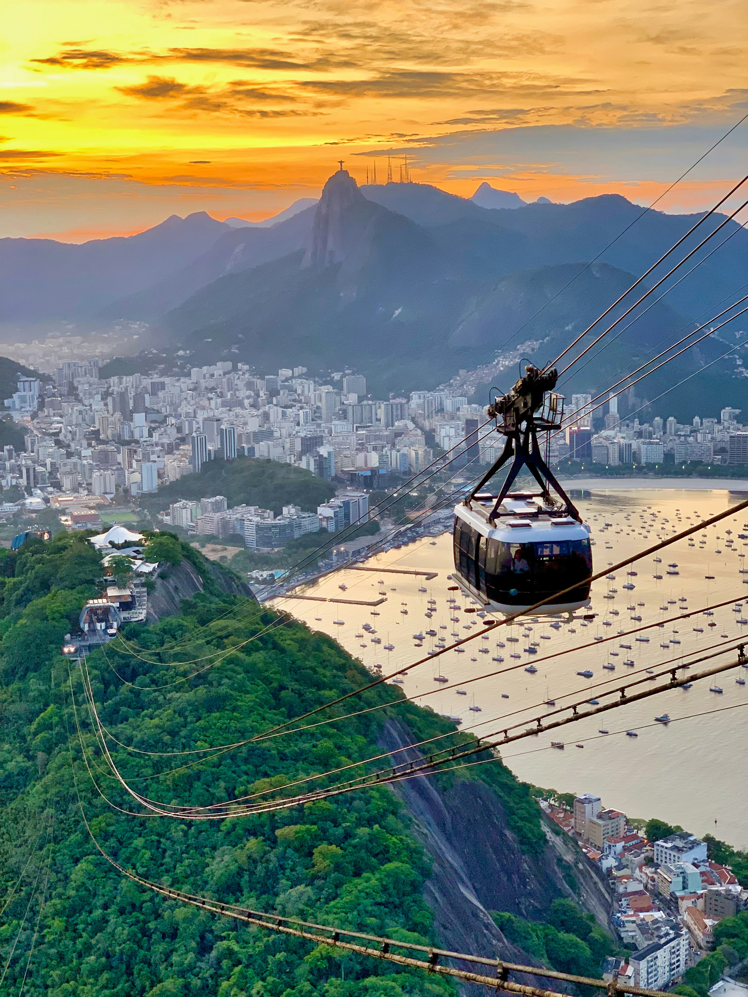 Cable car descending over a green mountain with cityscape and boats on water at sunset, Rio de Janeiro
