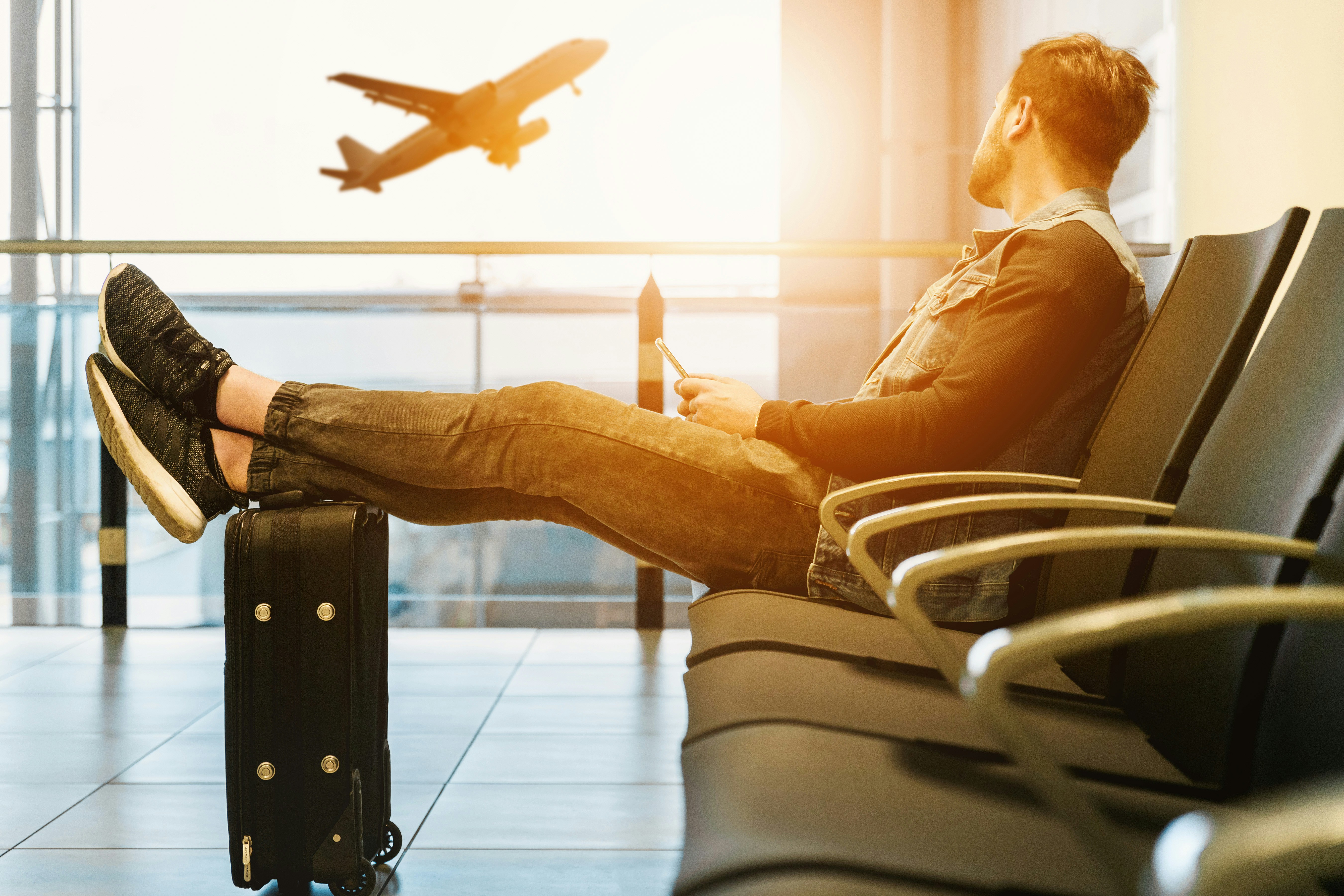 A man sitting in an airport waiting area with his legs resting on a suitcase, watching an airplane take off through large windows