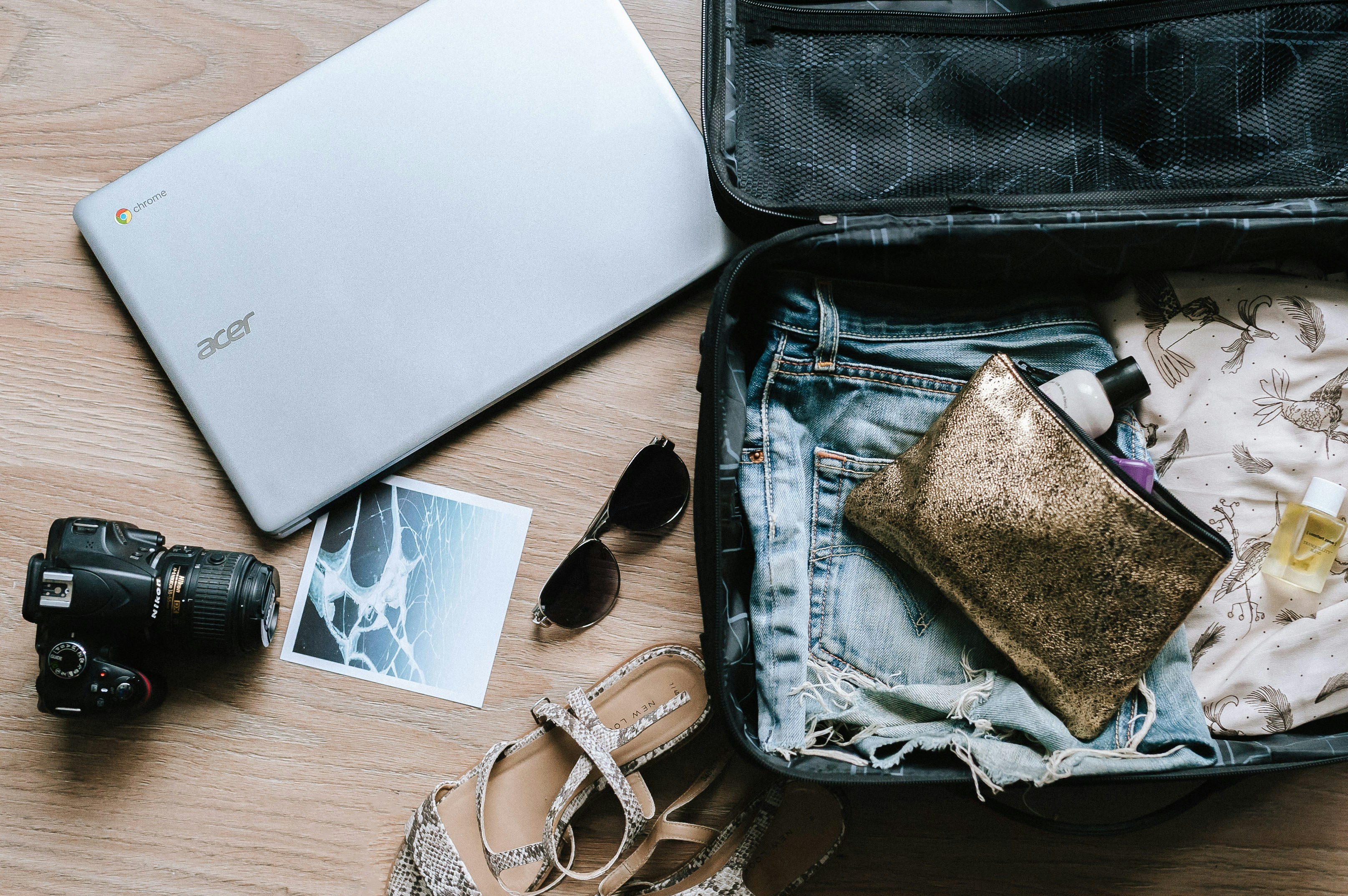 Partially packed suitcase with denim shorts, gold makeup bag, toiletries, and nearby items including laptop, camera, sunglasses, photo, and sandals on wooden floor