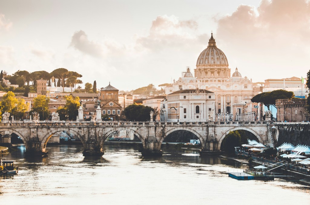 Stone bridge crossing river with statues and St. Peter's Basilica dome in background at sunset