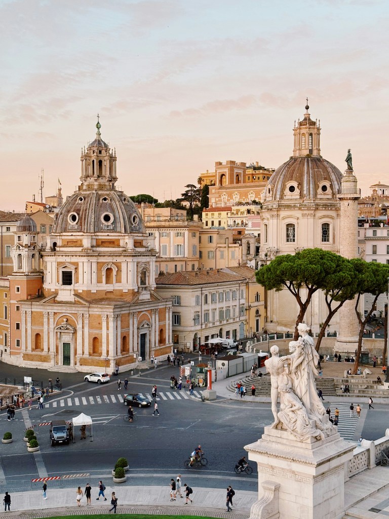 Historic city square with two large domed buildings, a tall column, and a white statue in the foreground at sunset